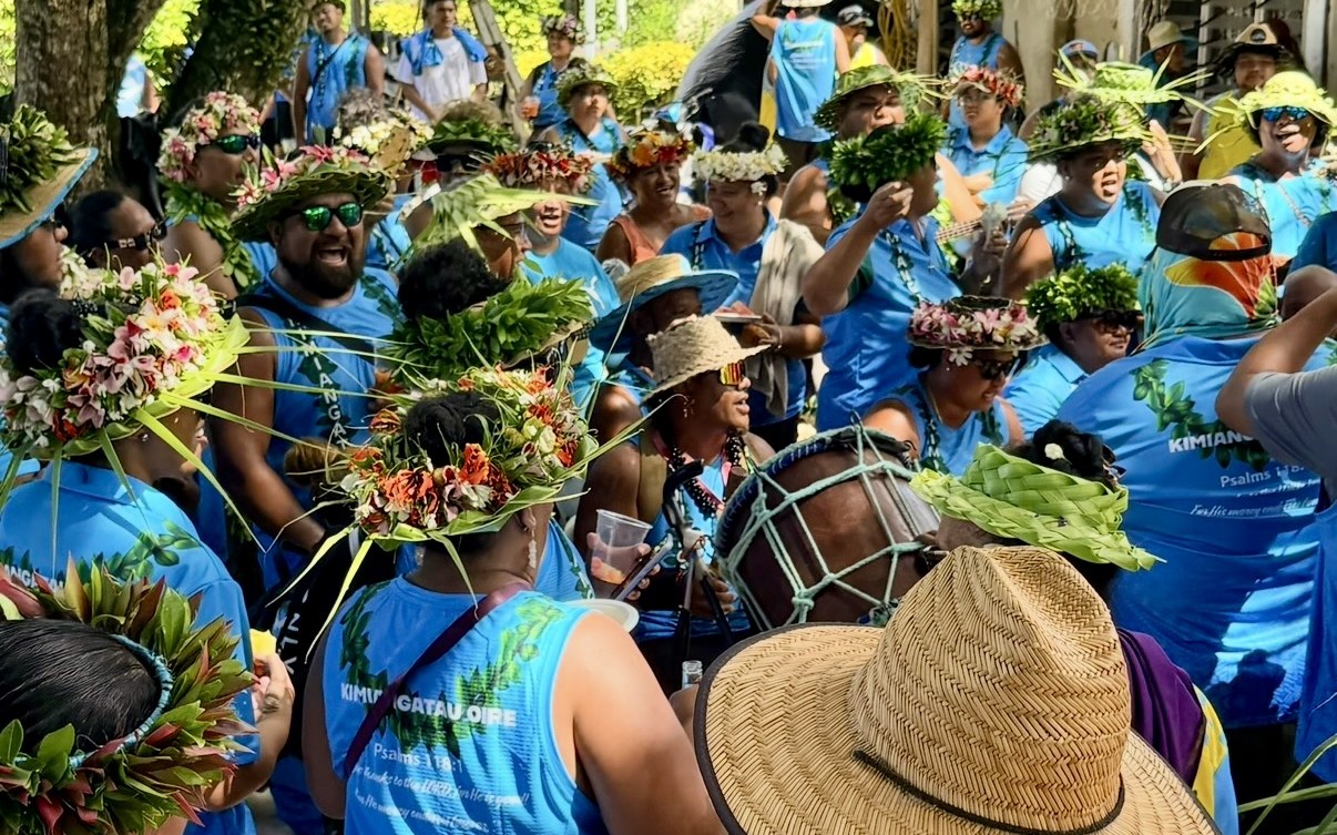 The people of Kimiangatau, Maʻuke, in blue, wearing ʻei katu of flowers and leaves, with a pate drum among them