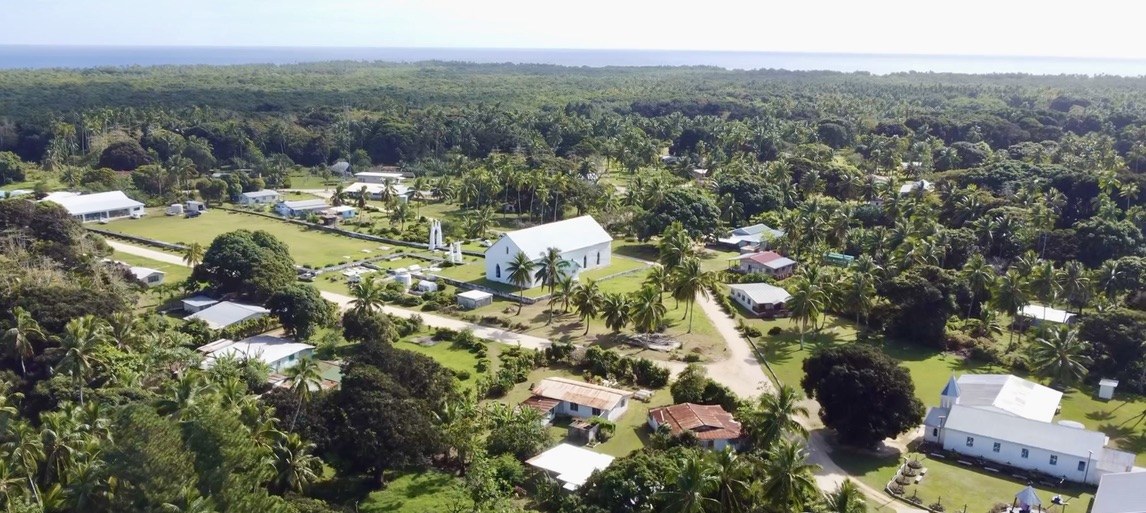 Oiretumu village on Maʻuke from the air &mdash; two white
              churches among the houses and palms, with the reef and the open
              sea along the horizon